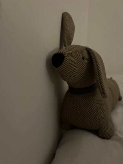 A stuffed dachshund toy is positioned leaning against a white wall in a dimly lit room. The toy has brown and beige herringbone fabric, a black nose, and floppy ears. A dark collar is visible around its neck. The toy rests on a white surface, possibly a bed or sofa. The background consists of the plain white wall and the edge of the room. The scene is static and devoid of human presence. The location is not discernible within the image itself.