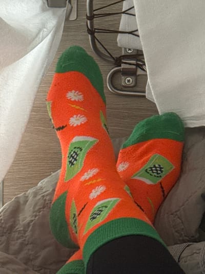 An indoor scene, depicting a pair of human feet, is observed in Gryazi, Russia. The feet are clad in matching bright orange socks with distinct green cuffs, heels, and toes. The socks feature a repeating pattern of white daisy-like flowers with yellow centers, thin black lines with yellow segments, and green geometric flag-like shapes containing a black hashtag symbol. Dark-colored trousers or leggings are visible just above the sock cuffs. The feet rest on a quilted grey fabric surface, likely a blanket or upholstery. In the background, a light brown surface exhibiting a wood grain texture is visible, along with sections of white fabric. A polished metallic fixture, possibly a bed frame component or a bracket, is also present in the upper right, secured by visible screws. The scene is evenly lit.