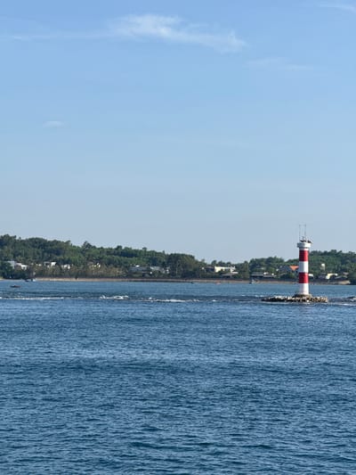 This image depicts a lighthouse in the waters off the coast of Tinh Khe, Vietnam. The lighthouse is striped red and white and stands on a rocky base in the middle of the blue water. In the background, a lush green coastline is visible, dotted with houses and trees. The sky is clear and blue with a few wispy clouds. The water is calm, with gentle ripples on its surface, and small waves are breaking around the base of the lighthouse. Several small boats are also visible in the distance, near the shore. The overall scene suggests a peaceful coastal environment.
