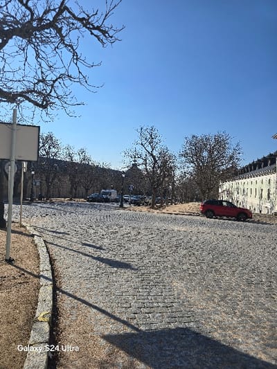 The image presents an outdoor scene in Real Sitio de San Ildefonso, Spain, under a clear, bright blue sky. A wide, grey cobblestone road, composed of interlocking rectangular pavers, curves from the lower left quadrant towards the right background. On the immediate left, a narrow strip of dry, light brown ground runs parallel to a stone curb that separates it from the cobblestone surface. Long, dark shadows, indicative of a low sun angle, stretch across this ground and a significant portion of the cobblestone road. A light-colored, vertical pole is visible in the left foreground.
In the midground, a multi-story building with a light-colored facade, numerous windows, and a dark, multi-pitched roof line the right side of the cobblestone road. A red SUV is parked parallel to this building on the cobblestones. Further into the background, a cluster of deciduous trees, predominantly bare of leaves, defines a central area. Beyond these trees, additional parked vehicles, including a white van, are visible on another road or parking surface. A building featuring a dark, dome-like structure can be faintly discerned among the trees in the central background. No individuals are present in the depicted scene. The bottom left corner of the image contains a "Galaxy S24 Ultra" watermark.
