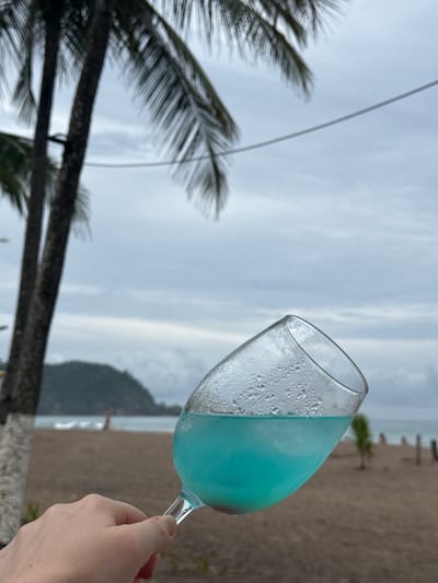 A close-up shot shows a hand holding a stemmed glass filled with a vibrant blue liquid, likely a cocktail. The glass is partially filled, with condensation visible on its exterior. In the background, a sandy beach stretches towards the ocean, with gentle waves breaking near the shore. A person is visible standing in the shallow water. A lush green hill forms the backdrop to the beach. Two palm trees stand in the foreground to the left, their fronds reaching towards an overcast sky. The scene appears to be set in Jacó, Costa Rica, a coastal town known for its beaches and palm trees.