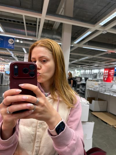 In an indoor commercial environment, a young woman with auburn hair is taking a selfie with her smartphone. She is wearing a light-colored corduroy jacket with pink trim and a pink hoodie. Her lips are pursed, and she has several silver rings on her fingers. A smartwatch with a pink band is visible on her left wrist. The background shows rows of shelves stocked with various items, typical of a large retail store such as IKEA, a common sight in many cities, including Son en Breugel, Netherlands. The lighting suggests fluorescent overhead fixtures, and signage is visible in the distance.
