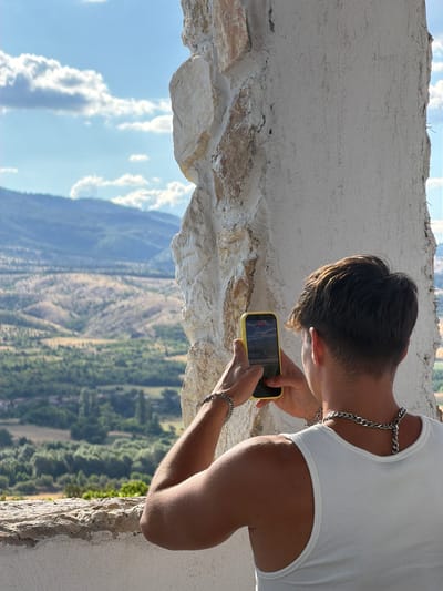 The image depicts a person, from behind, wearing a white ribbed tank top and a silver chain necklace, holding a yellow smartphone to capture a scenic view. The person's hair is dark and styled in a way that suggests a modern haircut. They are standing on a stone structure, possibly a balcony or a lookout point, with a rough, textured wall on their right and a ledge in the foreground. The background reveals a vast landscape of rolling hills covered in lush green trees and drier, lighter vegetation, with distant mountains under a bright blue sky dotted with fluffy white clouds. The lighting suggests it is daytime, likely midday or afternoon. The overall scene conveys a sense of travel and appreciation for natural beauty.
Based on the context provided, this image was taken in the vicinity of Kocherinovo, Bulgaria. The location appears to be an elevated vantage point offering a panoramic view of the surrounding countryside. The architectural elements, such as the rough stone and plastered walls, could be part of an older structure or a specifically designed viewpoint in or around Kocherinovo. The natural landscape, with its distinct hills and vegetation, is characteristic of the Bulgarian terrain.