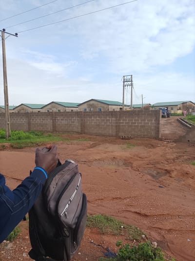 In Kopowabam, Nigeria, a person's arm and shoulder are visible in the foreground, holding a dark grey backpack with zippers and pockets. The backpack has white lettering. The person is wearing a dark blue jacket with white stitching on the sleeves and a blue stripe around the cuff. The scene depicts a construction site or developing residential area. In the background, a low wall constructed of concrete blocks runs horizontally. Beyond the wall, a row of residential buildings with green roofs is visible. A utility pole with electrical wires stands to the left of the frame. Further in the background, more utility poles and a transformer are visible. A white truck is parked on a dirt path to the right, and a person in pink clothing is standing near the truck. The ground is covered in reddish-brown dirt with some patchy green grass. Several small puddles are present on the ground, indicating recent rainfall. The sky is overcast with scattered clouds.