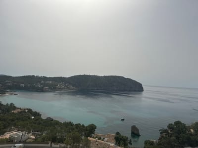 The image depicts a coastal scene in Andratx, Spain, characterized by a bay surrounded by land formations and vegetation. The sky is overcast.
A prominent landmass, covered in trees and houses, borders the bay on the left side of the frame. Across the bay is a mountainous landform, likely a peninsula, also covered with dense vegetation.
In the bay, the water transitions in color from turquoise near the shore to a deeper blue further out. A small motorboat is visible on the water's surface. In the lower right of the image, a rocky outcropping juts from the water near the shoreline. The foreground features trees and the rooftops of buildings, indicating an elevated viewpoint overlooking the bay.