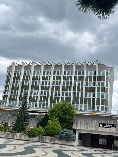 This media file shows a cloudy day in Créteil, France, featuring a modern building with a facade of numerous windows and a distinctive sawtooth-like concrete overhang. The building appears to be an administrative or office building, with signage visible that reads "C.C.A.S. SERVICE DES SENIORS" (a social services department for seniors) and "LA POSTE" (the French postal service). A large, decorative tiled pathway with wavy, abstract patterns is visible in the foreground, suggesting a public space. Lush green trees and shrubs surround the building, adding a touch of nature to the urban setting. The sky is overcast, indicating a possibly cool or rainy day. There are no people visible in the image. The overall scene suggests an urban environment in France, likely a public service area or a business district.