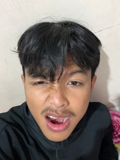 A close-up, angled photograph shows a person's face with a scrunched expression. The person has dark hair styled forward, dark eyes, and a mustache. Their mouth is slightly open, revealing some teeth and tongue. The person is wearing a black top. The background appears to be a textured, light-colored wall. The location is in the city of Paweden, Indonesia.
