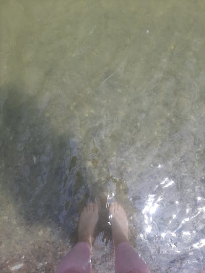 The image shows a person's bare feet submerged in shallow, murky water, with sunlight glinting off the surface. The water appears to be on a pebbly shore, with some darker, possibly sandy areas visible. The person is wearing pink pants that extend up to their ankles. The scene is likely a natural body of water, such as a riverbank or lake shore, in Sukholuzhzhia, Ukraine. The water's clarity is low, obscuring the bottom in most areas, though some rocks and debris are visible. The light suggests it is daytime.