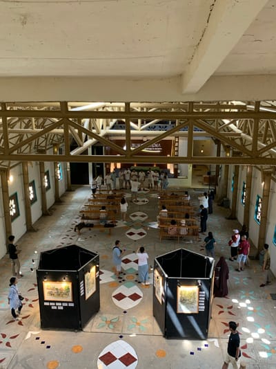 The image shows a large, open, indoor space with a tiled floor featuring a geometric design in shades of brown, yellow, blue, and white. The space is located at the Borobudur Temple, a UNESCO World Heritage site in the Kecamatan Borobudur district of Magelang Regency, Central Java, Indonesia. The space is viewed from above. The image shows a group of people standing in a circle in the middle of the room, while others are walking around or looking at artwork displayed on black hexagonal stands. Two large black hexagonal structures stand facing each other in the center of the room, each showcasing a painting. The ceiling is made of exposed wooden beams. The overall feeling of the space is one of light and openness.