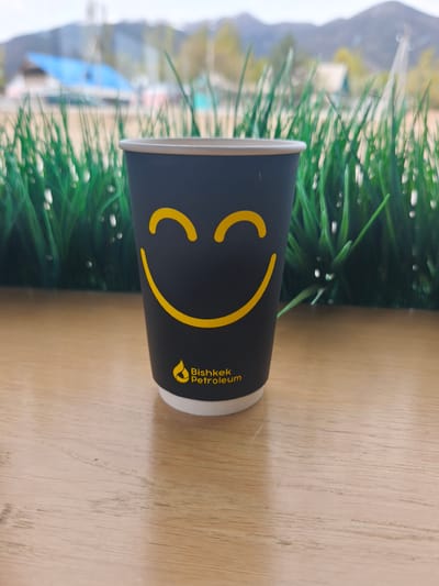 A close-up shot shows a black paper cup with a yellow smiley face and the words "Bishkek Petroleum" and a logo printed on it. The cup sits on a light brown wooden surface, possibly a table. In the blurred background, there's a view of green grass-like plants, buildings with a blue roof, and mountains under a slightly overcast sky. The image seems to be taken in an outdoor setting, most likely in the city of Bishkek, Kyrgyzstan, based on the branding on the cup.