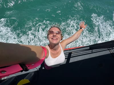 A woman with blonde hair and sunglasses takes a selfie while standing on a boat with turquoise water and white wakes. She is wearing a white tank top and pink shorts, with a pink strap over her left shoulder. Her right arm is raised and outstretched. The boat's railing and deck are visible in the foreground, with shadows cast on the deck. The scene is outdoors, under bright sunlight.