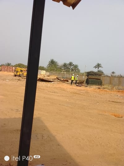 The image depicts an outdoor industrial or storage yard in Hoche, Nigeria, under a light, hazy sky. A dark, vertical structural column partially obstructs the left foreground, with a corrugated roof edge visible at the top. The ground is unpaved, reddish-brown dirt, covering most of the lower half of the frame.

In the midground, two individuals are standing on the dirt. Both wear yellow-green high-visibility vests and white hard hats. They are positioned near a concrete perimeter wall that extends horizontally across the scene, topped with what appears to be barbed or razor wire. To the immediate right of the individuals, a large stack of bundled metal mesh or rebar is present. To their left, scattered debris, including what resembles wood and metal fragments, lies on the ground. Further to the left in the midground, a yellow mobile crane truck with an extendable boom is parked. Behind the crane truck, several stacked red-brown shipping containers are visible. A light pole with a fixture is situated behind the individuals, near the concrete wall.

The background features numerous palm trees and other green vegetation behind the wall and containers. The "itel P40 4G" watermark is present in the lower-left corner. No direct interaction between the individuals or specific actions are clearly discernible; they appear to be observing or standing within the yard.