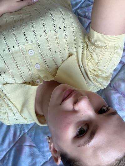 The image presents a close-up, high-angle perspective of a young adult, appearing female, lying down indoors. The subject is wearing a pale yellow, short-sleeved knit top featuring an openwork or eyelet pattern with vertical columns of small perforations and scattered larger holes. The garment has a ribbed texture on the collar, button placket, and sleeve cuffs, with three white, pearl-like buttons fastened on the front. A manicured hand with short, light-colored nails is partially visible near the upper left side of the torso.

The subject's face is tilted slightly, with direct eye contact towards the camera. Features include fair skin, dark eyes with visible eyelashes, and well-defined eyebrows. The lips are slightly parted, exhibiting a natural pink hue. A small, light-colored stud earring with a gold-tone setting is visible on the earlobe. Dark hair is visible at the very bottom edge of the frame. The subject is positioned on a quilted surface, likely a blanket or bedspread, which displays varying shades of soft blue, purple, and grey, arranged in a stitched diamond or square pattern. The scene is observed indoors, reportedly located in Nizhnyaya Gar', Russia.