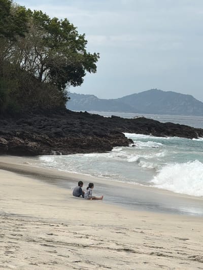 Two young children are playing on a sandy beach next to the ocean. The child closer to the camera is sitting up, wearing a dark blue shirt. The child further away is lying down, wearing a light-colored shirt with a floral pattern. The ocean waves are crashing on the shore, creating white foam. In the background, dark, rocky cliffs rise from the water, and beyond that, hazy mountains are visible. Lush green trees and vegetation are present on the left side of the image, with their branches extending towards the sky. The sky is overcast and light gray, suggesting a cloudy or hazy day. The sand on the beach is light tan and shows footprints. The scene appears to be a serene and natural coastal environment. The time of day is likely daytime, given the lighting. There is no visible text in the image.