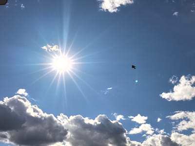 A bright sun with distinct rays shines in a clear blue sky, partially obscured by fluffy white and gray clouds. The sun's glare is intense, creating a starburst effect. In the mid-ground, a bird is captured in flight, silhouetted against the sky. The lower portion of the image features dramatic, shadowed clouds that contrast with the bright sunlit ones above. The scene suggests a daytime setting with varied cloud cover, typical of fair weather. There is no visible text in the image.