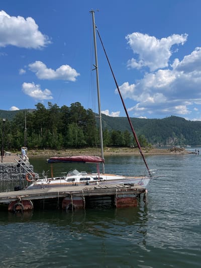 This image captures a serene scene at a dock, likely in Divnogorsk, Russia. A sailboat, with the name "Julia" visible on its side, is moored to a wooden pier. The boat is white with orange accents and has a prominent mast. The water surrounding the dock is calm and reflects the clear blue sky dotted with fluffy white clouds.
In the background, a densely forested hillside rises steeply from the shore, its green expanse meeting the clear blue sky. The time of day appears to be midday, given the bright sunlight casting shadows and the vibrant colors. The weather is fair, with a gentle breeze suggested by the rippling water.
On the dock, a person wearing a blue shirt is visible, seemingly engaged with the boat or its surroundings. A life preserver is also on the dock, a common safety feature at a marina. The dock itself is made of wood and is lined with rocky material on one side, possibly for erosion control or as a foundation.
Visible text on the boat includes "Julia" and "P18-13K4". The overall mood of the image is peaceful and tranquil, evoking a sense of leisure and connection with nature. The setting suggests a popular spot for sailing and outdoor activities on the water.