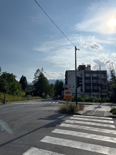 The image shows a street intersection in Bled, Slovenia, at the coordinates 46.368 latitude and 14.113 longitude. A single cyclist is riding on the left side of the road towards a crosswalk. There are trees and greenery on either side of the road, with a large concrete building with "Hotel Park" on the side on the right.  In the background, there are mountains. There are three traffic signs: a white sign pointing to the right with "Bled - Prekmurska cesta," an orange sign pointing to the right with "Pristani sve Bled castle," and a white sign with a red circle and black diagonal line inside. There is a streetlight and power lines overhead. The sky is blue and slightly cloudy. The sun is partially visible in the upper right corner of the image.