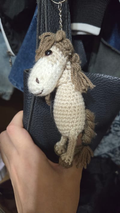 A close-up, high-angle shot shows a crocheted horse keychain being held in a person's hand. The keychain is light brown and cream-colored with yarn for its mane and tail. It has black bead eyes and a small brown stitched nose. The horse is lying on top of a black textured surface, likely a purse or bag, which is itself resting on some blue fabric, possibly denim. The background is blurred and consists of dark colors. The lighting appears to be indoor artificial light. The image does not contain any visible text. The scene is static and focuses on the detail of the handmade item.