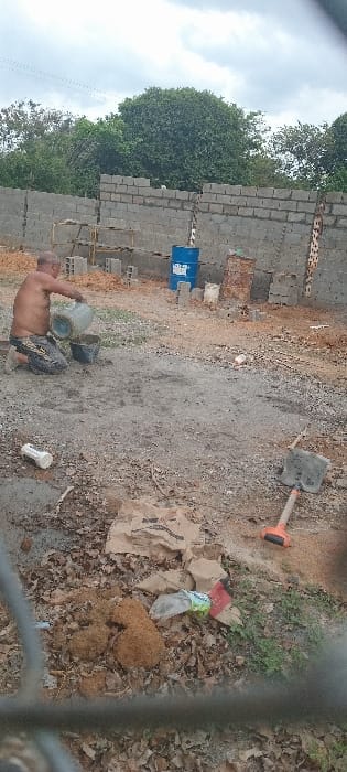 A shirtless man carries two buckets on a dirt lot with a cinder block wall in the background. A blue barrel and other items are stacked against the wall. Debris litters the ground, including leaves, cardboard, and an orange-handled shovel. The scene is viewed through a blurred foreground, possibly a fence.