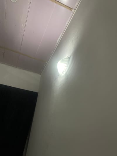 The image shows a close-up of a wall with a light fixture. The light is on and casts a glow on the wall. The wall appears to be painted a light color and has some texture. Above the wall, part of a ceiling is visible, which appears to be made of panels with lines dividing them. There is also a dark area in the lower left of the image, possibly a doorway or a piece of furniture covered in dark fabric. The scene is indoors, and the lighting suggests it is nighttime or a dimly lit environment. No people are visible in the image. The location is identified as being in Pantinapu, Nigeria.