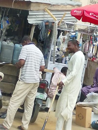 The image displays an outdoor street or market scene in Babban Giji, Nigeria. Four adult males are visible, interacting within the environment. In the left foreground, a man with his back mostly to the viewer wears a horizontal white and blue striped polo shirt and light-colored trousers, looking downwards at an object in his hands. A black motorcycle with a visible license plate, RBP 589QR, is positioned behind him.

To the right, a man in a long, light-colored (off-white/cream) traditional garment stands in profile, facing left, with an open-mouthed expression suggesting laughter or speech. He holds a wooden stick, resembling a cue stick, with its tip angled towards the ground. Small red or orange ornaments are visible in his hair.

In the background, a commercial stall or shop with a corrugated metal roof is present. The left portion of the stall features multiple grey and blue plastic jerrycans, coiled blue cables, and various hanging merchandise. The right portion of the stall is an open display containing numerous small items such as white PVC pipe fittings, plastic containers, small fans, and other hardware. A red umbrella branded "Fresh" provides shade over the right side of this stall. A partial sign displaying "GIDAN ST" is visible on a light-colored wall above the right side of the stall.

Two additional males are partially visible: one seated behind the laughing man, wearing a light pink shirt and leaning forward; another seated on the far right, wearing a dark purple garment, with only his lower body visible. The ground surface appears to be unpaved.