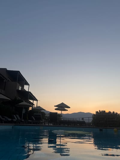 This media file depicts an outdoor scene at dusk. The primary subjects include a swimming pool with reflections of surrounding trees and buildings, and a modern hotel or resort building with multiple balconies and open-air corridors. The sky transitions from a light blue to a warm orange-yellow near the horizon, indicating sunset. In the background, silhouetted mountains are visible, suggesting a coastal or mountainous landscape.

Several objects are present, such as lounge chairs, umbrellas, and a yellow ball floating in the pool. The overall setting appears to be a vacation resort, likely in Agios Nikolaos Municipal Unit, Greece, given the location context. There are no people visible in the frame.

The time of day is clearly sunset, characterized by the soft, warm light and the gradient in the sky. The weather appears to be calm and clear. The scene evokes a sense of tranquility and relaxation, often associated with evening at a resort. No visible text is present in the image. The image is oriented vertically, suggesting it was captured with a phone camera.