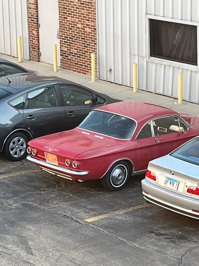 The image captures a parking scene in Woodridge, Illinois, United States. A red vintage car is parked next to a modern gray sedan and a tan BMW. The vehicles are in an exterior setting of a parking lot with yellow line markers on a gray concrete surface. The background features a building exterior with alternating sections of red brick and white metal siding, including a window. Yellow bollards are spaced intermittently along the building's base. No people are visible in the photograph.