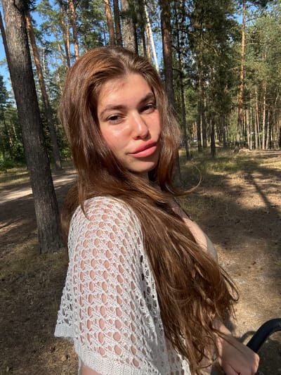 This media file shows a close-up portrait of a young person with long, light brown hair, lying down outdoors. The subject is facing the camera, with their head tilted to the side. Their eyes are open, and they appear to be looking directly at the viewer. Their lips are slightly parted, and they are wearing light pink lipstick. They are wearing a white, crocheted top that reveals glimpses of their skin through the open-knit pattern. The background is a forest, with tall pine trees and dry, grassy ground. Sunlight filters through the trees, casting dappled shadows. The overall impression is one of a relaxed and natural moment.

The subject's expression could be interpreted as calm or perhaps a little wistful. The lighting suggests it is daytime, likely a sunny afternoon, given the warmth of the light and the shadows. The presence of pine trees and dry, low vegetation is consistent with a forest environment in a temperate climate. The specific location of Kurovskoye, Russia, can be inferred, contributing to the context of a natural, wooded area. There is no visible text in the media file.
