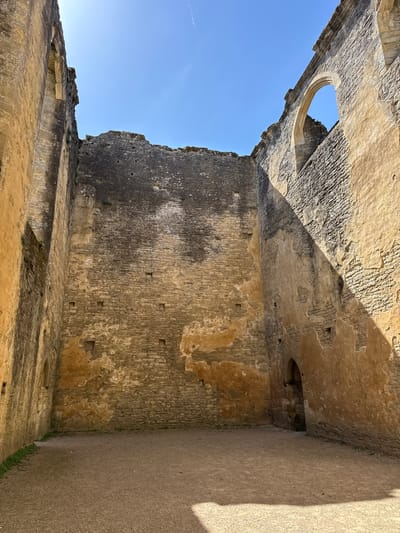 The image shows the interior of Minster Lovell Hall in West Oxfordshire, United Kingdom. The walls are made of rough-hewn stone and are in a state of disrepair. There are gaps and holes in the walls, and the surface is weathered and stained. A round window is visible in the upper left corner, through which the blue sky can be seen. The ground is covered in dirt. The image is taken from a low angle, looking up at the walls and sky.