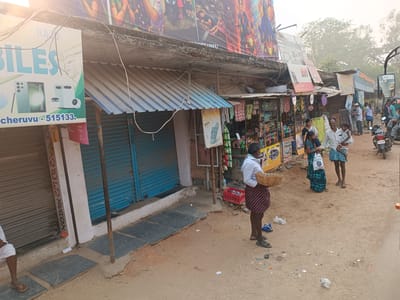 The image captures a street scene in Kothacheruvu, India. The foreground features a partially visible man in a white shirt and maroon skirt, holding a woven basket. To his left, shops with closed blue shutters line the street, partially obscured by signage and awnings. Further down the street, open-front shops display goods, and people are gathered, including a man holding a child. A parked motorcycle is visible on the street. The street itself is dirt-covered with scattered debris. Buildings and advertisements are stacked on top of each other. The sky is bright, contributing to a slightly hazy atmosphere.