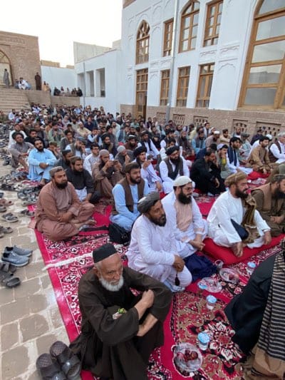An outdoor gathering of approximately 150 adult males is depicted, seated on red and patterned rugs laid on a paved ground. The individuals are predominantly of South Asian descent, dressed in traditional clothing, including various styles of shalwar kameez and turbans. Many have beards. The group is oriented towards a large, white building with ornate arched windows and decorative white molding, likely a historical structure or a religious institution. To the left of the building, a staircase with a stone balustrade leads up to an entrance, and a few people are visible on the stairs. Scattered around the periphery of the gathering are numerous pairs of sandals and shoes. The lighting suggests it is late afternoon or early evening. The presence of food and drink items, such as dates and small cups, on the rugs indicates a communal meal or refreshment. The scene appears to be in a public outdoor space within the city of Kandahar, Afghanistan, given the architectural style and attire of the individuals.