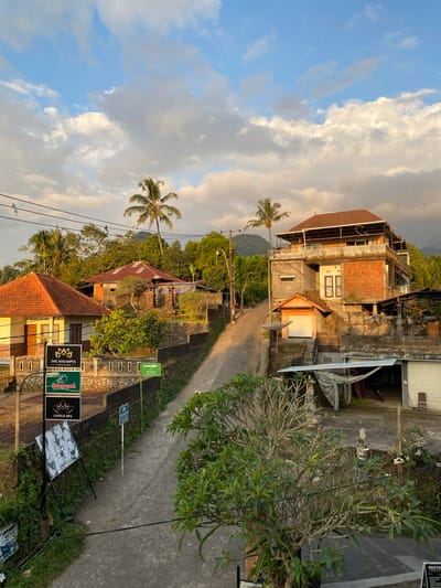 A narrow, paved road winds uphill through a village in Sawan, Indonesia, flanked by traditional Balinese houses and lush tropical vegetation. The golden hour light casts long shadows and illuminates the scene with a warm glow. Palm trees are scattered throughout the landscape, with a prominent one on the left and another to the right of a multi-story building.

On the left side of the road, a yellow house with an orange tile roof sits behind a low stone wall. Adjacent to it, a series of dark green signs are mounted on poles, indicating "THE SEKUMPUL VILLA & SPA", "Sekumpul Restaurant & Coffee", and "LOTUS SPA". Further up the road, a taller building constructed from brick and concrete dominates the right side. It features multiple balconies and a brown tiled roof. In front of this building, a smaller structure with an orange roll-up door is visible.

The environment is characterized by a hilly terrain with dense greenery. The sky above is a blend of blue with scattered white and gray clouds, catching the warm tones of the setting sun. Power lines crisscross the upper portion of the image, and a distant mountain range is visible in the background, shrouded in a light haze. The scene depicts a peaceful rural setting in a tropical climate.