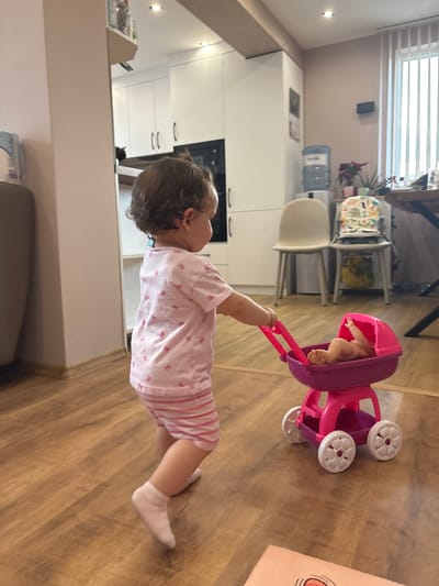The image shows a young child playing with a pink toy stroller indoors. The child, seen from the side, is wearing a pink and white striped outfit and white socks. The stroller contains a doll. The setting appears to be a kitchen or living area with wooden flooring. In the background, white kitchen cabinets and a water cooler are visible. The scene is brightly lit, suggesting daytime. The overall mood is one of playful engagement. There is no visible text in the image. The location context is Strelcha, Bulgaria.