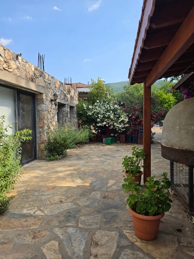 This image depicts an outdoor courtyard with a stone-paved floor. To the left, there is a building with a stone wall and a large glass door. In front of the building, lush green plants are growing. Beyond the building, a dense cluster of trees with white and pink flowers is visible, interspersed with stacks of colorful plastic crates. To the right, a wooden-framed roof extends over the courtyard, supported by a wooden post. Beneath the roof, a large, domed structure, possibly a pizza oven or a similar cooking appliance, is situated on a metal stand with wheels. In the foreground on the right, two terracotta pots contain green potted plants. The background features a hilly landscape under a clear blue sky with scattered clouds. The scene suggests a rustic, possibly rural or semi-rural setting, consistent with areas outside the immediate city center of Bodrum, Turkey, potentially in a residential or guesthouse environment with an outdoor cooking area. There are no people or animals present in the image, and no specific actions or interactions are depicted.