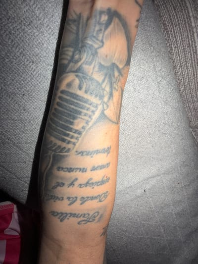 A forearm displays a tattoo with a microphone image accompanied by Spanish text that reads "Familia Donde la vida espiga y el amor nunca termina". The skin tone of the arm is light. The arm rests on a gray textured fabric.