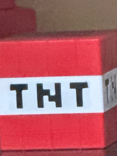 A close-up, eye-level shot shows the side of a red block with a white stripe bearing the black pixelated letters "TNT." The block is viewed from the front left, with the right edge of the block angled away from the camera. The lighting is even, with no harsh shadows or highlights. The background is blurred and consists of muted brown and red tones, suggesting an indoor setting or a textured surface. This object is located in Heniivka, Ukraine.