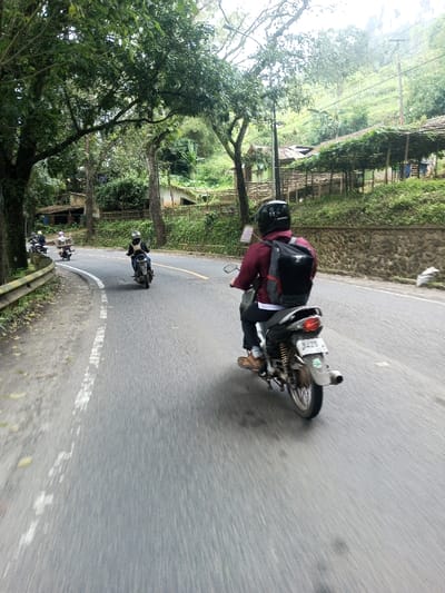 An image captured from a moving perspective depicts a multi-lane asphalt road curving to the left, likely within the city of Khajuripada, India. In the immediate foreground, a person wearing a dark helmet, a maroon long-sleeved shirt, dark trousers, and brown shoes rides a dark-colored motorcycle. A black backpack with gray accents is worn. The motorcycle's rear license plate partially displays "8475." Further along the curved road, two additional motorcyclists are visible, also proceeding in the same direction. The first wears a helmet and dark attire, while the second, further back, transports a large, light-colored package or box on their motorcycle.
To the left of the road, a dirt shoulder with a white dashed line transitions to a metal guardrail. Beyond this, large trees with dense green foliage line the road. To the right, a low stone wall runs parallel to the road, topped with green vegetation. Behind this wall, the terrain ascends into a lush, green hillside. Scattered along the hillside are several simple structures, including houses with light-colored exteriors and dark roofs, and a prominent covered structure with a lattice-like roof, possibly for cultivation. Utility poles with overhead wires are also present on the hillside. The background features heavily vegetated hills under an overcast sky, indicating daylight conditions.