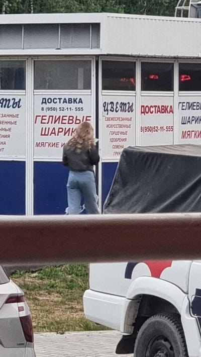 The image shows a storefront with several windows. On the left, text reads " ДОСТАВКА" and "ГЕЛИЕВЫЕ ШАРИКИ". In the center, text reads "ЦВЕТЫ" and "ДОСТАВКА". On the right, text reads "ГЕЛИЕВЫЕ ШАРИКИ". A woman with blonde hair is standing in front of the window, facing away from the camera, and appears to be holding a phone. She is wearing a dark jacket and light blue jeans. Behind her, a black tarp-covered object is visible. In the foreground, a portion of a white vehicle is visible. The storefront appears to be located in an urban or suburban area, with a grassy area and trees in the background. There are no specific landmarks or indications in the image to pinpoint the exact location within Megion, Russia.