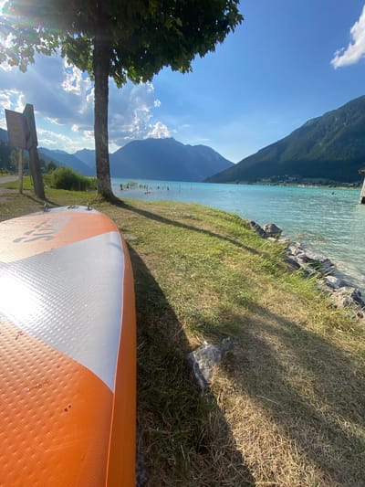 This is a media file depicting an outdoor scene on a sunny day at Lärchenwiese, Austria. The camera angle is low and tilted, giving a perspective from the ground. In the foreground, the tail end of an orange and white paddleboard is visible, resting on dry grass. Beyond the paddleboard, a grassy area slopes down towards a body of clear, turquoise water. The water is calm, with visible rocks and a few small buoys or markers in the distance.

The background features a magnificent mountainous landscape under a bright blue sky dotted with fluffy white clouds. The mountains are covered in lush green vegetation. A tree trunk stands vertically in the left middle ground, with its branches and leaves providing shade. Further in the distance, along the shoreline of the lake, a small village or cluster of buildings can be observed.

The overall atmosphere is serene and suggests a peaceful day by the water, likely during summer given the lush greenery and bright sunshine. The lighting indicates it is daytime, with the sun casting shadows on the grass. There is no discernible activity of people directly visible, but the presence of the paddleboard implies recreational use of the lake. No significant text is clearly legible in the image.