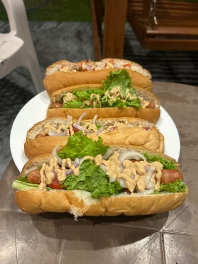A platter of four loaded hot dogs is presented on a table. The hot dogs are arranged in a staggered, layered fashion on a white plate, with the front-most hot dog closest to the viewer. Each hot dog features a split bun, a sausage, lettuce, chopped tomatoes, sliced red onions, and a drizzle of a creamy, beige-colored sauce. The background of the image reveals an outdoor setting with a glimpse of a white chair on the left and wooden swing seating on the right. The ground appears to be paved. The scene is likely taking place in Kadawatta, Sri Lanka, given the contextual information.