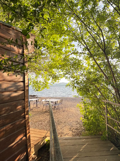 The image shows a wooden structure in the foreground, with a textured metal roof. Beyond the structure, a wooden deck with a metal railing leads to a pebbled area adjacent to a body of water. Several metal chairs and tables are set up on the deck, suggesting an outdoor seating area. Overhanging trees with green leaves provide shade and frame the scene. The water appears calm, with a slight ripple, and a hazy landscape is visible in the distance. The sunlight suggests it is daytime, possibly late afternoon due to the warm tones and shadows. The overall impression is a serene, natural setting, perhaps a lakeside cafe or resting spot. There is no visible text.