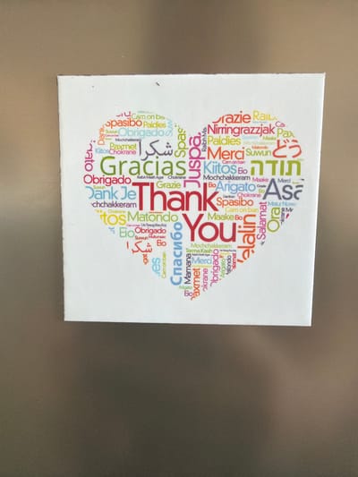 The image shows a square canvas art piece displayed on a textured, dark surface, possibly a refrigerator. The canvas features a heart-shaped word cloud with various expressions of "thank you" in multiple languages, such as "Gracias," "Merci," "Dank Je," "Obrigado," "Arigato," "Grazie," "Suwun," "Paxmet," "Mochchakkkeram," "Siyabonga," "Spasibo," "Raiba," "Nirringrazzjak," and others. The words are arranged in different colors and fonts, with "Thank You" being the most prominent phrase in the center. The art piece appears to be a decorative item, possibly a magnet or a small plaque, placed on a surface. There are no people or discernible elements of the city of Sderot, Israel, in this image.