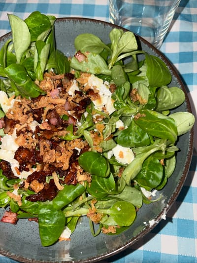 A close-up, top-down shot captures a vibrant salad served in a dark grey or blue ceramic bowl. The main components of the salad are fresh, bright green lamb's lettuce (corn salad), generously mixed with crumbled white cheese, possibly feta or goat cheese. Numerous pieces of dark, caramelized bacon or pancetta are scattered throughout, alongside lighter, pinker bacon chunks. Golden-brown, crispy fried onions or shallots provide an additional textural element, topping the entire dish.
The bowl has a rustic, textured finish with a slightly irregular rim. It rests on a table covered with a blue and white checkered tablecloth, which fills the background. A clear glass, partially visible in the upper left corner, is positioned beside the bowl.
The lighting is soft and even, suggesting an indoor setting. There are no people visible, and the scene depicts a prepared meal, ready for consumption. No specific time of day is discernible, and no text is visible within the image. The overall impression is of a fresh and hearty salad, likely part of a meal in a casual dining environment in Urbanización Encinares De Alcolea, Spain.