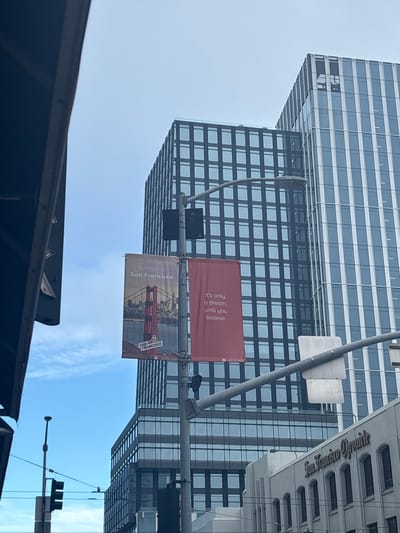 In this urban scene in San Francisco, California, United States of America, two banners hang from a lamppost in front of modern glass-facade skyscrapers. The left banner features an image of the Golden Gate Bridge with the text "Believe in San Francisco" and "SAN FRANCISCO" in the lower portion. The right banner, a solid red, displays white text that reads "It's only a dream, until you believe." To the right of the skyscrapers, a building with the prominent signage "San Francisco Chronicle" is visible, indicating a downtown or financial district location. Traffic lights and street wires are present, suggesting a street-level view. The sky is overcast with some blue visible.