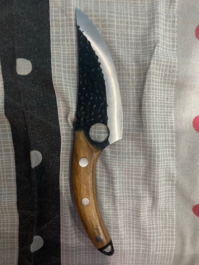 The image displays a fixed-blade knife resting vertically on a patterned fabric surface. The knife features a recurve blade with a polished, sharp edge and a contrasting textured, dark-colored spine. A circular cut-out is present near the ricasso. The handle is made of light brown wood, secured by three silver-colored pins, and includes a dark lanyard hole at its pommel. The fabric background exhibits two primary patterns: a vertical dark grey and light grey stripe pattern with irregular light grey ovular shapes on the left, and a finer light grey grid pattern on a white background on the right, which also shows a partial red circular element in the lower right. The specific location within the city of Borovan, Bulgaria, cannot be ascertained from the visual evidence presented in this image.