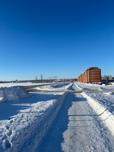 A winter scene shows a long, straight road covered in snow, flanked by snow-covered terrain. A tall, brick building sits along the road. Power lines stretch into the distance, parallel to train tracks. The sky is a clear, bright blue. Shadows of trees and poles fall across the snowy landscape.