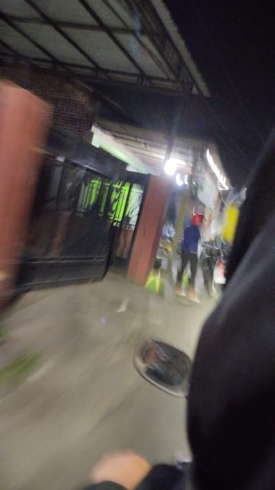 The image, taken at night in Bangsri, Indonesia, depicts a blurry, motion-filled scene, likely from the perspective of someone riding a motorcycle. In the foreground, a dark sleeve and a hand are visible. A motorcycle's side mirror reflects a partial view of the rider and the surrounding environment. The middle ground shows two individuals. One, wearing a blue jacket and a red helmet, stands facing away from the camera. The other person, partially obscured and also appearing to be moving, is next to them. Behind them, a dimly lit structure with a metal roof and brickwork is visible, alongside a large, dark object that might be a stall or a vehicle. Green vertical elements, possibly fencing or bars, are present on a structure to the left. The ground appears to be paved, and lighting fixtures are intermittently visible in the background, suggesting a street or market setting. The overall impression is one of rapid movement and low light conditions.