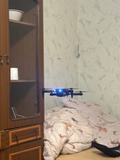 The media file displays an indoor scene, possibly a bedroom, in Vilnius, Lithuania. The main subjects are a small black drone with blue LED lights, hovering mid-air, and a bed covered with a white duvet featuring a pattern of small, reddish-brown animal silhouettes. To the left of the drone, the wall appears to have a light green textured wallpaper with faint, abstract patterns. Below the drone and wall is a wooden piece of furniture, likely a cabinet or dresser, with a reflective surface and visible drawer handles. The lighting suggests it could be daytime, as there is natural light, but the exact time of day is unclear. The overall impression is a casual, domestic setting, with the drone appearing to be in operation within the room. There is no visible text in the image.