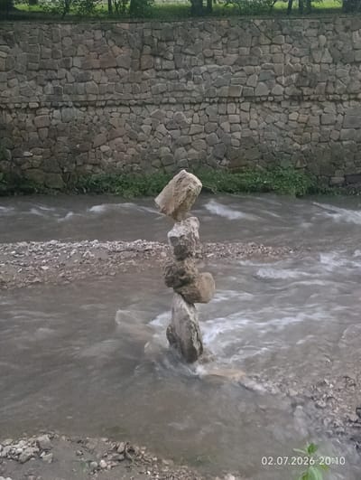 A stone wall forms the backdrop of a scene featuring a river and a stacked rock formation. The wall, constructed of irregularly shaped, light-colored stones, runs horizontally across the upper portion of the image. Below the wall, the river flows, its water appearing murky and dynamic with visible white streaks indicating movement. The riverbed is composed of small stones and gravel. A precarious stack of five stones stands in the middle of the river, the largest at the bottom, diminishing in size towards the top. The top stone is a rough, angular piece. On the left and right banks of the river, a strip of green grass and some vegetation is visible, with trees suggested at the very top of the image. The overall lighting suggests it is either late evening or an overcast day. In the bottom right corner, a timestamp "02.07.2026 20:10" is visible, indicating the date and time. The scene appears to be located in Kislovodsk, Russia.