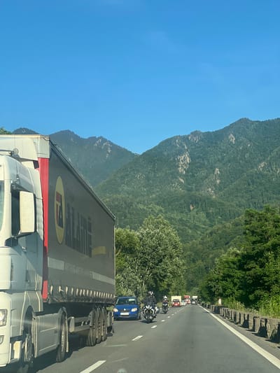 The image shows a view from a vehicle on a highway in Brezoi, Romania, with mountains in the background. A large white semi-truck with a dark grey trailer, bearing a yellow and red logo, is in the left foreground. The truck has several axles and wheels visible. To the right of the truck, a blue car is in the same lane. Further down the highway, there are several motorcycles with riders wearing helmets, and other vehicles including trucks and cars. The highway is asphalt with a white line marking the lane. On the right side of the road, there is a stone barrier. The surrounding environment consists of lush green trees and dense forests covering the mountains. The sky is clear and blue, suggesting a sunny day.